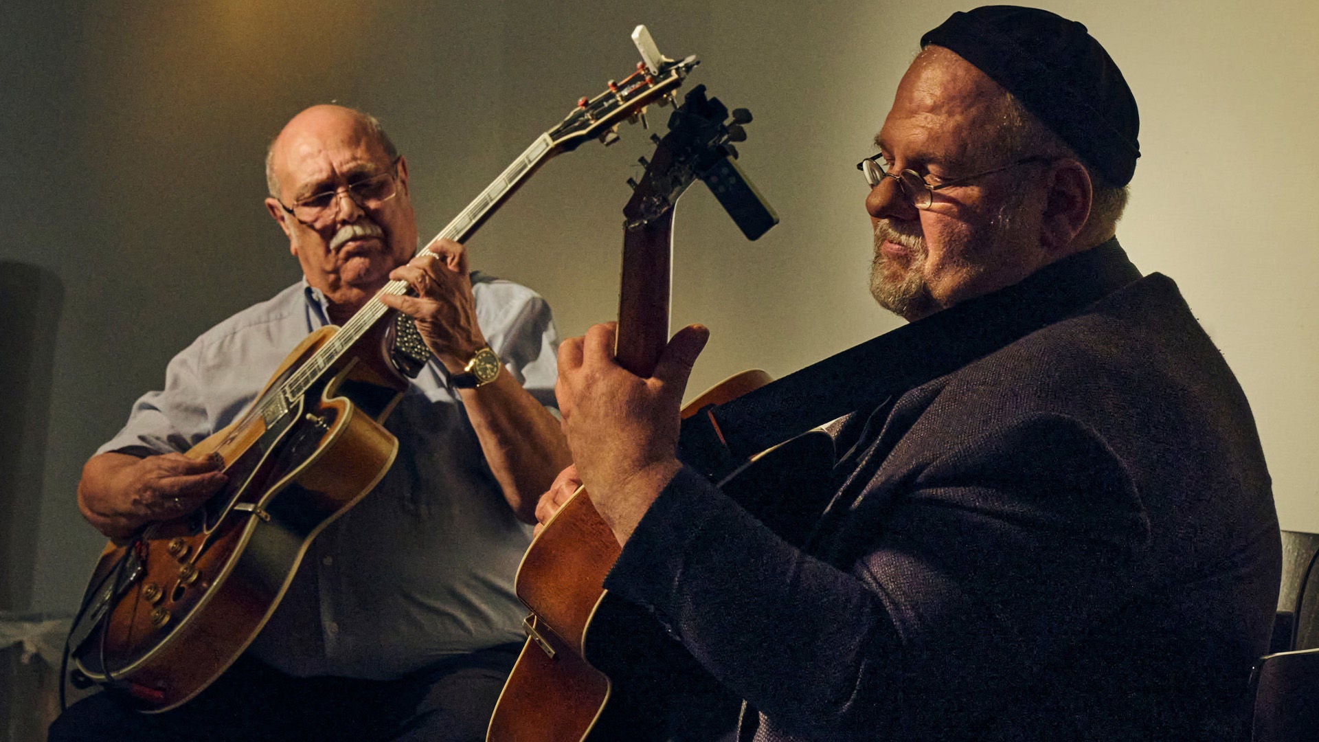 Zwei ältere Männer spielen akustische Gitarren in warmem Bühnenlicht; beide wirken konzentriert, sitzen einander zugewandt und erzeugen eine intime Konzertatmosphäre.