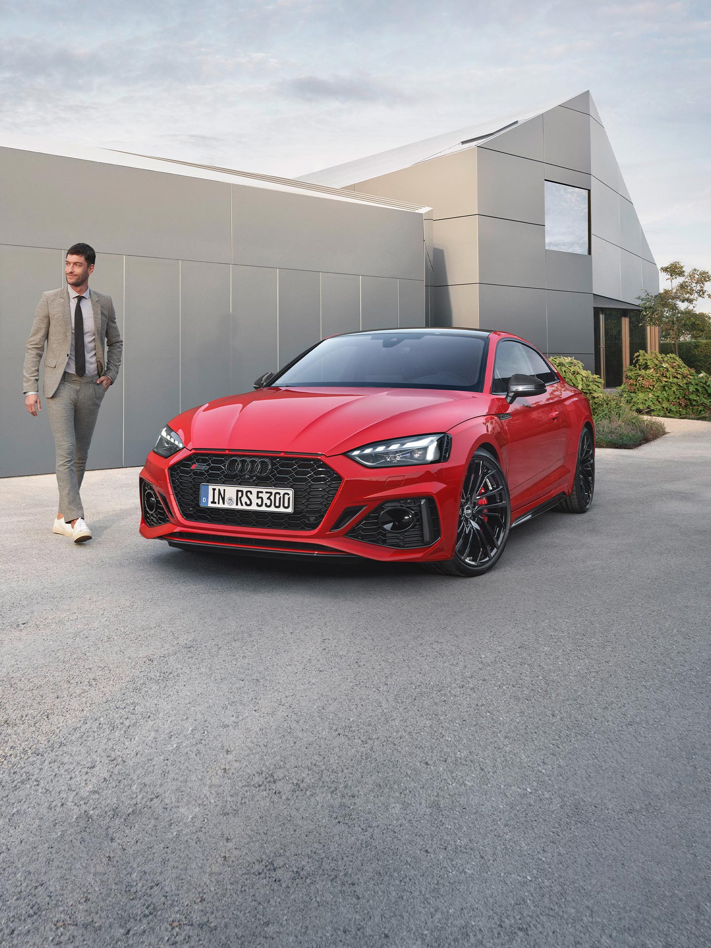 A red Audi RS5 Coupe parked outside a modern building with a person approaching it.