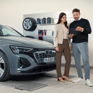 Mann zeigt Frau etwas auf einem Tablet neben einem grauen Audi in einer Werkstatt mit Felgen und Zubehör an der Wand.