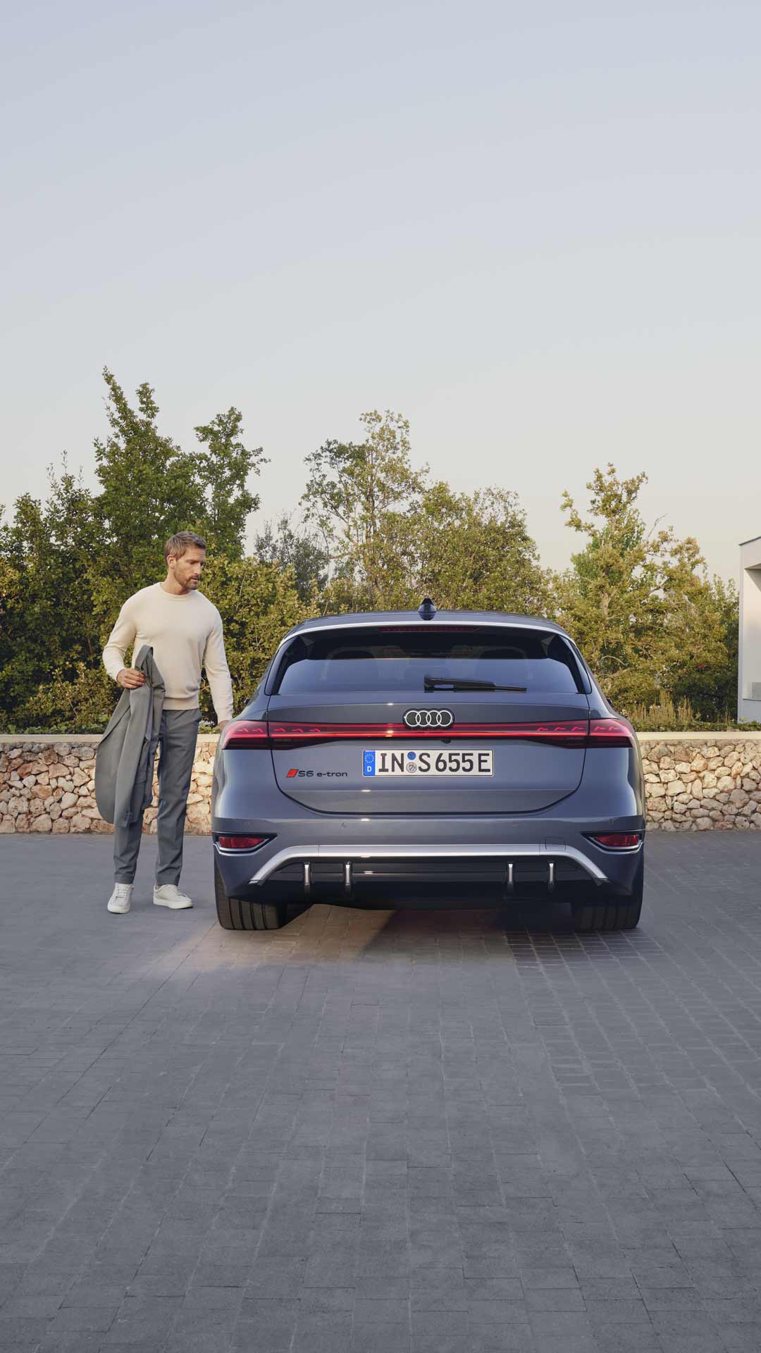 Rückansicht eines grauen Audi. An der Fahrertür steht ein blonder Mann mit einer grauen Jacke in der Hand. Er scheint kurz davor, in das Auto einzusteigen.      