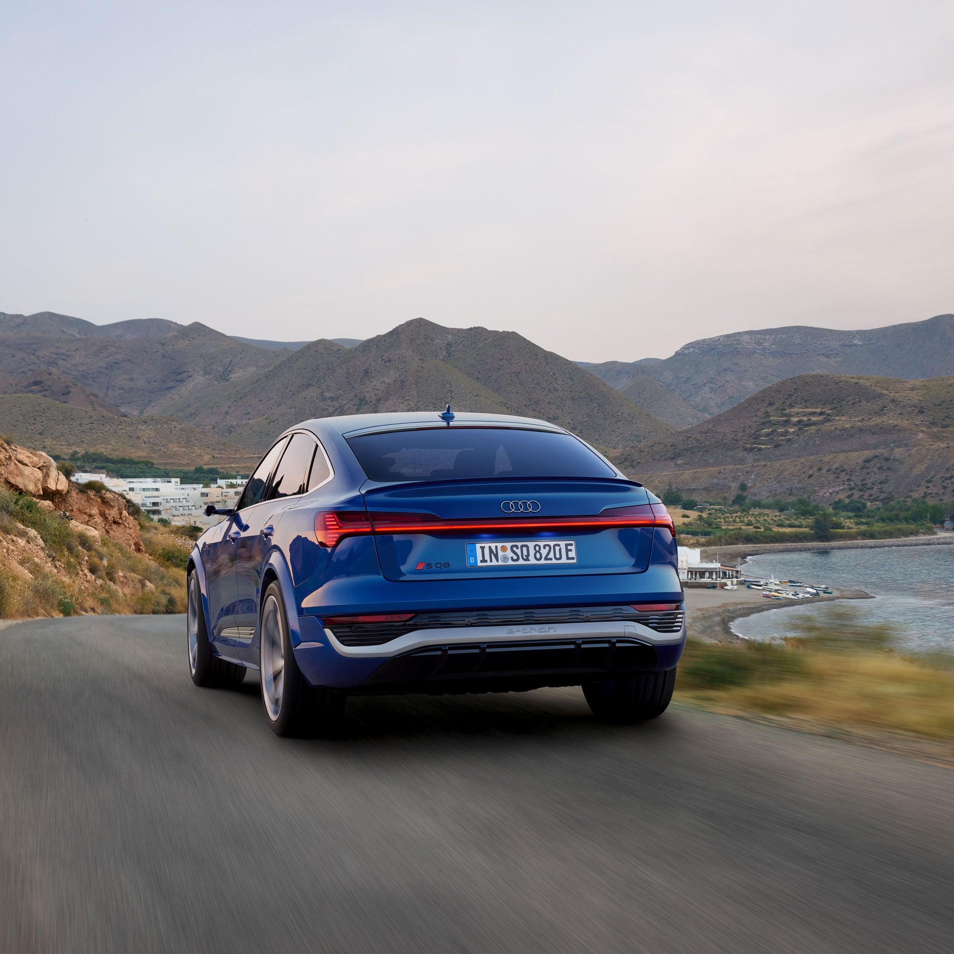 Ein blauer Audi fährt auf einer Straße am Wasser im Hintergrund sieht man eine bergige Landschaft.