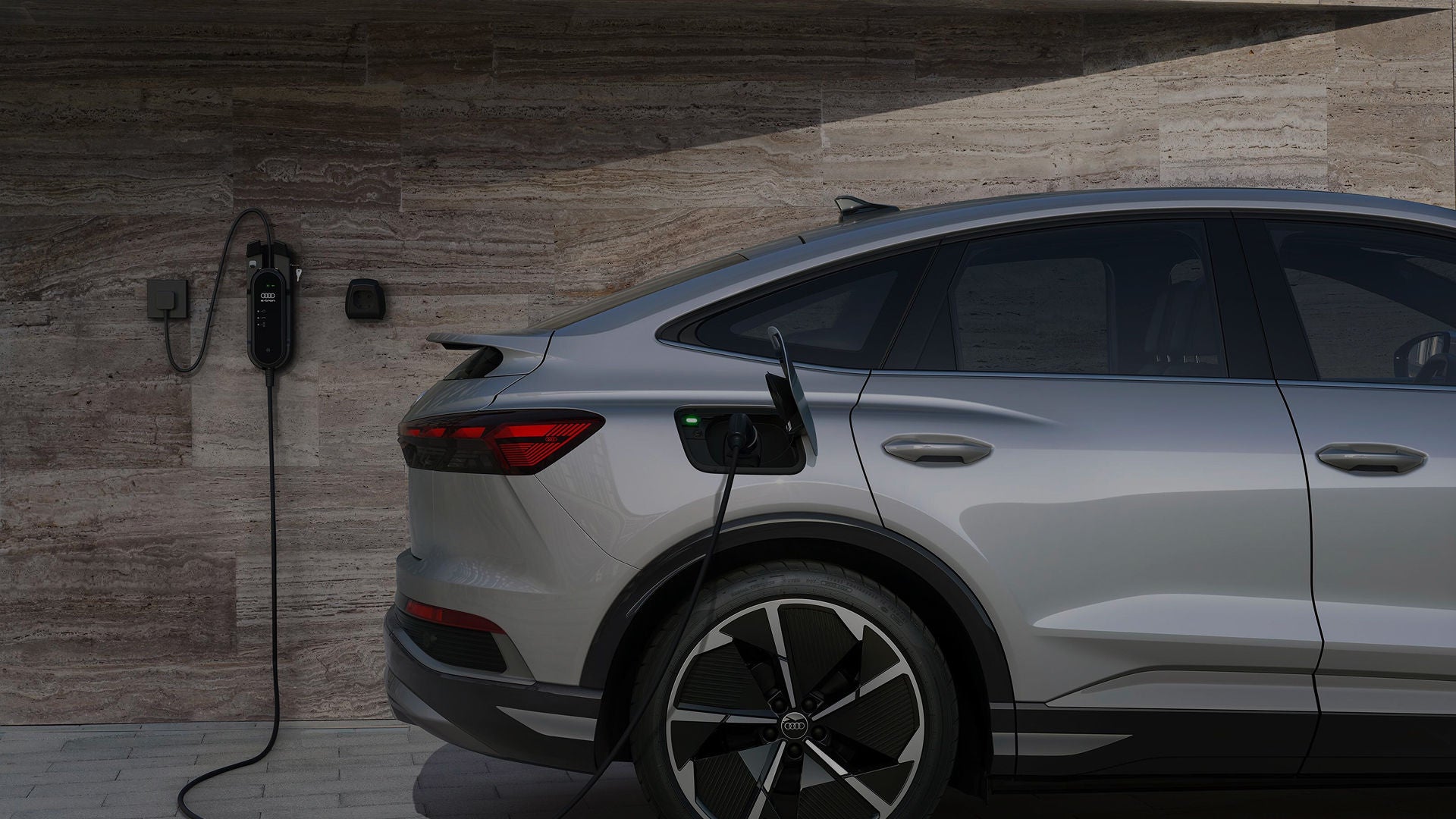 A silver Q4 Sportback e-tron plugged into a wall-mounted charger, set against a textured stone wall backdrop, showcasing modern design.