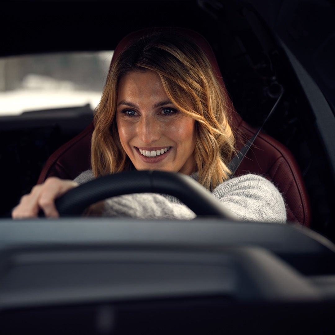 A smiling woman is sitting behind the wheel of a car, looking straight ahead.