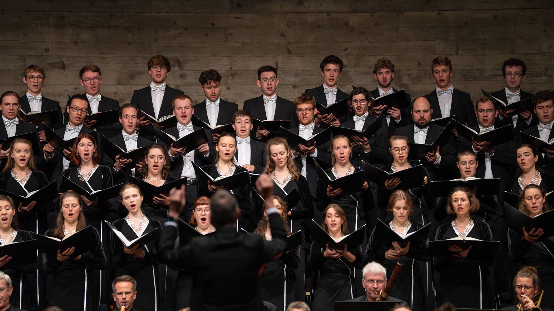Großer Chor in schwarzer Konzertkleidung singt auf der Bühne; davor dirigiert ein Mann.