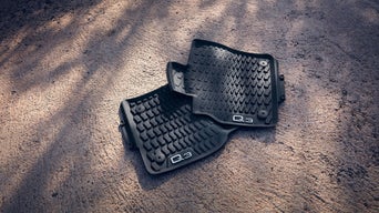 Zwei schwarze Audi Q3-Autofußmatten liegen auf hellem Boden, aufgenommen von oben.