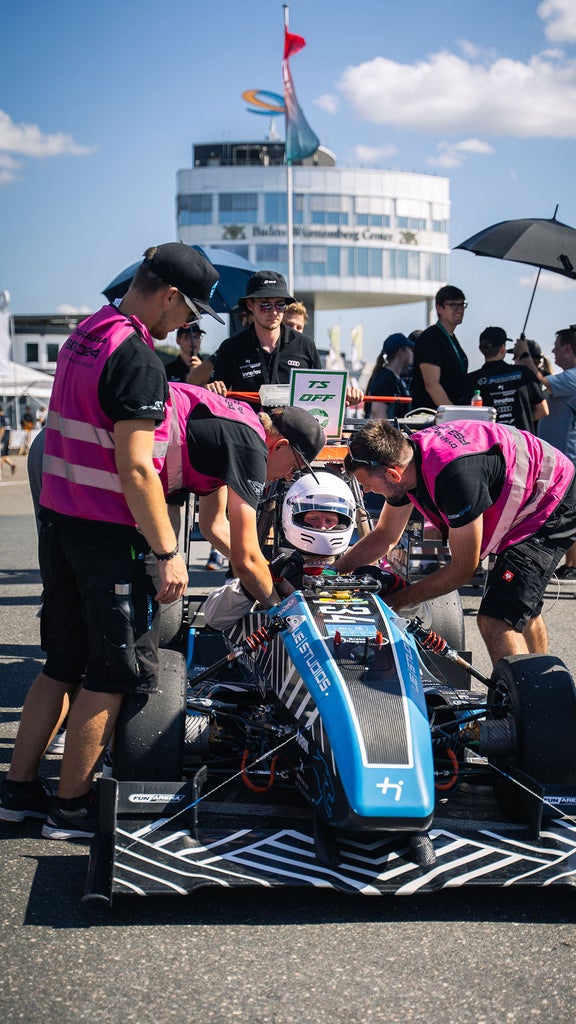 Ein Fahrer sitzt mit Helm in einem Rennauto und andere Personen stehen um das Auto herum und arbeiten daran