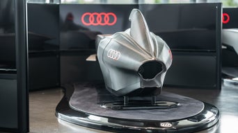 Silberner Audi racing Simulator mit hoher Rückenflosse auf einer runden Plattform vor schwarzen Wänden mit roten, leuchtenden Audi Ringen.