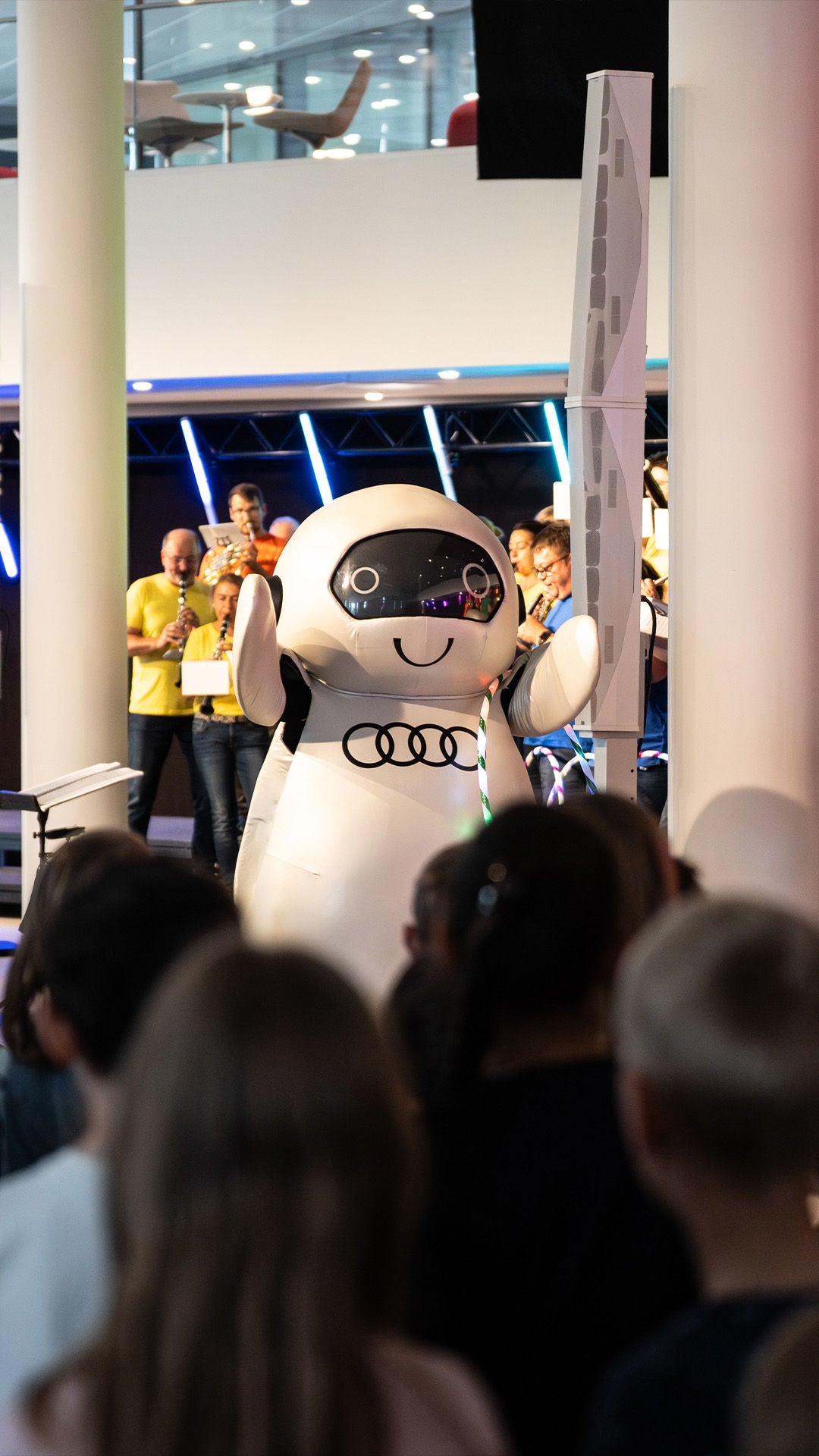 Maskottchen in weißem Audi-Kostüm steht vor Publikum; dahinter Musiker mit Blasinstrumenten in heller Innenhalle.