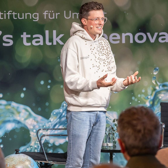 A man stands on a stage in front of an audience and gesticulates with a microphone. In the background: “Let's talk Greenovation”.