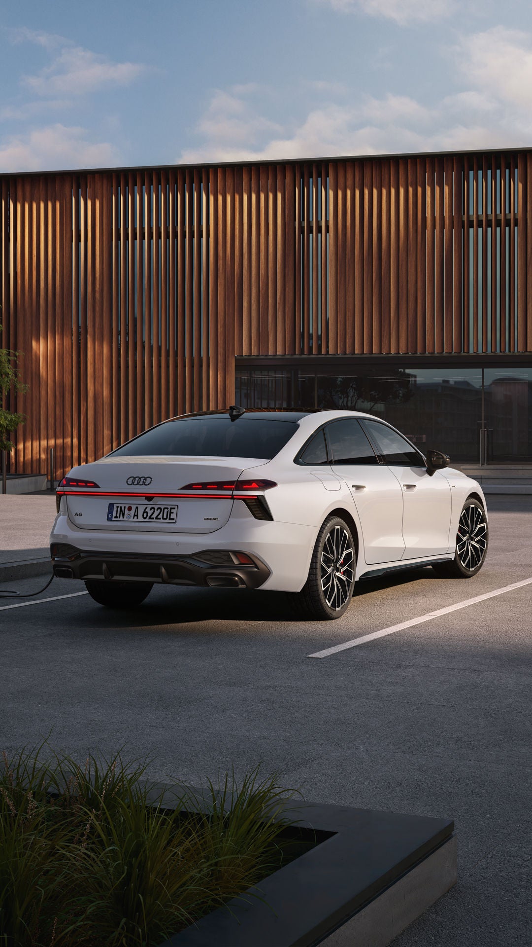 Die A6 Limousine e-hybrid in weiß steht auf einem Parkplatz vor einer Gebäudefassade. Eine junge Frau schließt die A6 Limousine e-hybrid an eine Ladestation an.