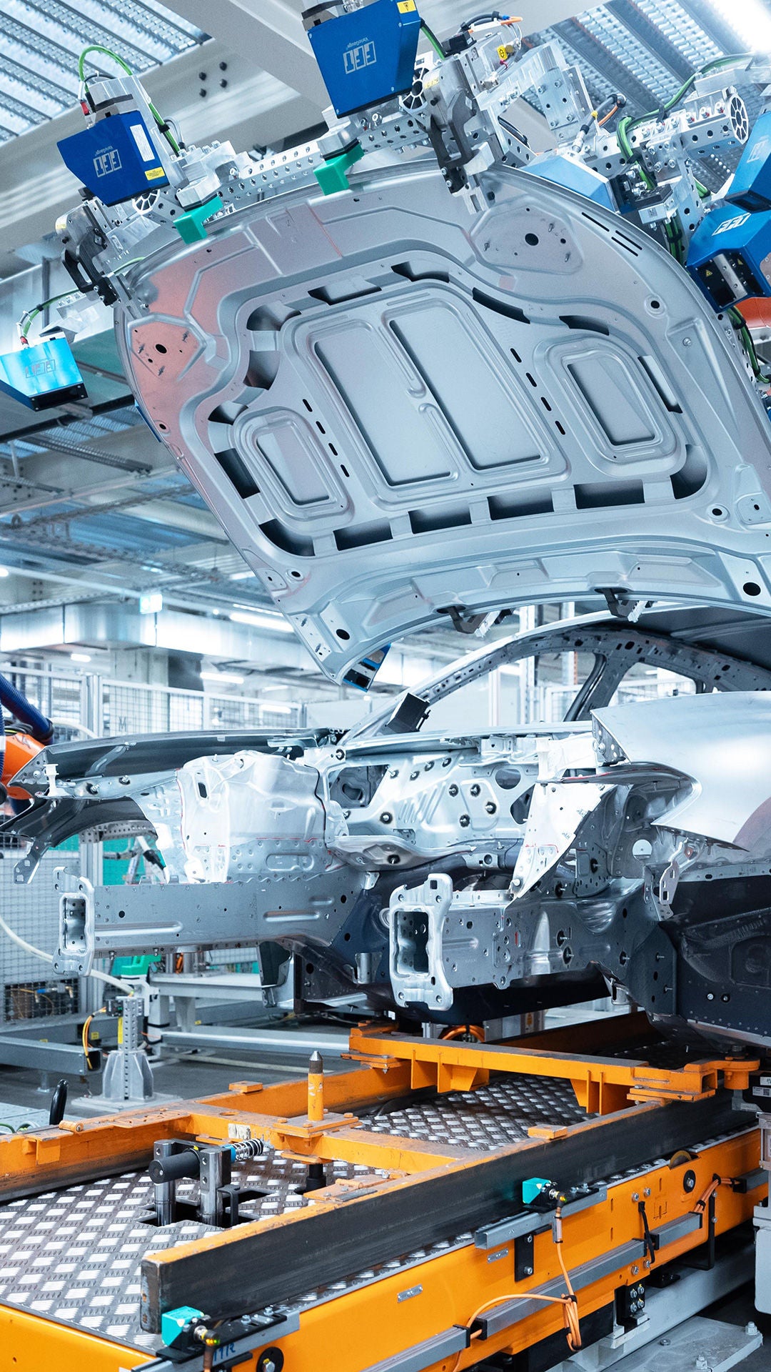 A modern production robot in a production hall lifts the body of a car. Surrounded by industrial machinery and lighting, the image shows a high-tech assembly process.