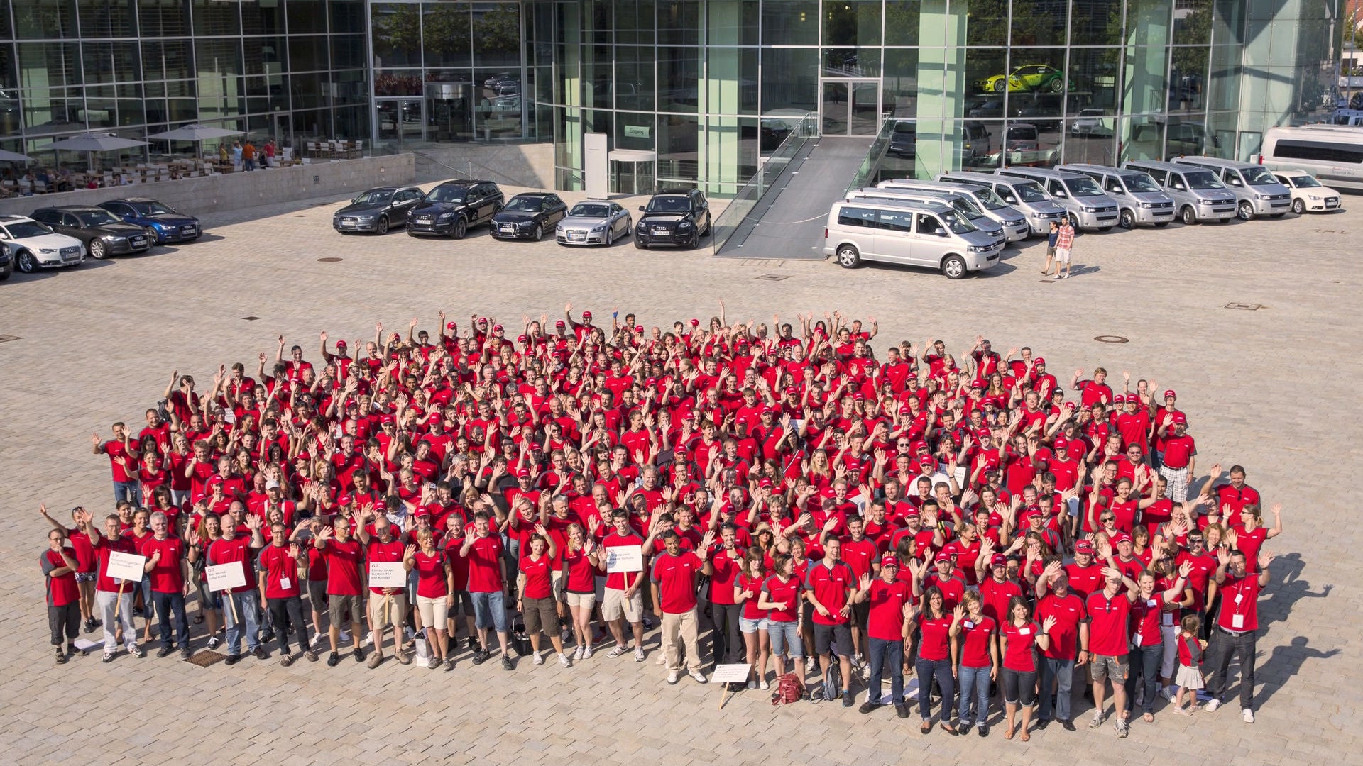 Luftaufnahme einer großen Gruppe von Audi Mitarbeitenden in roten Shirts. Sie stehen vor dem Firmensitz und winken in die Kamera.