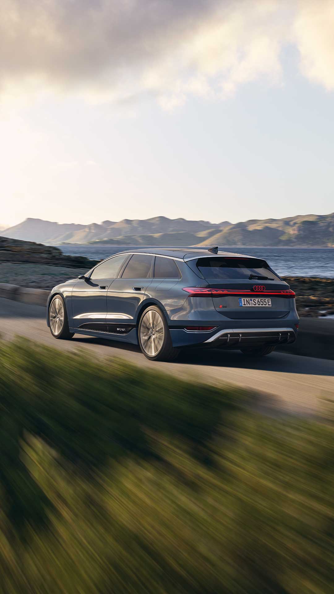 Dunkelblauer Audi S6 Avant e-tron fährt an einer Küstenstraße entlang, im Hintergrund Berge und Meer bei Abendlicht.