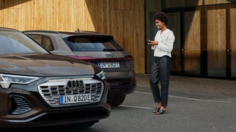 Frau mit Smartphone steht neben zwei geparkten Audi Q8 e-tron vor einem Gebäude mit Holzfassade.          