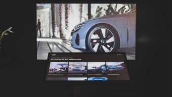 Großbildschirm zeigt Audi-Fahrzeug, davor interaktiver Touchscreen mit Menü zu Audi-Diensten auf dunkler Fläche.