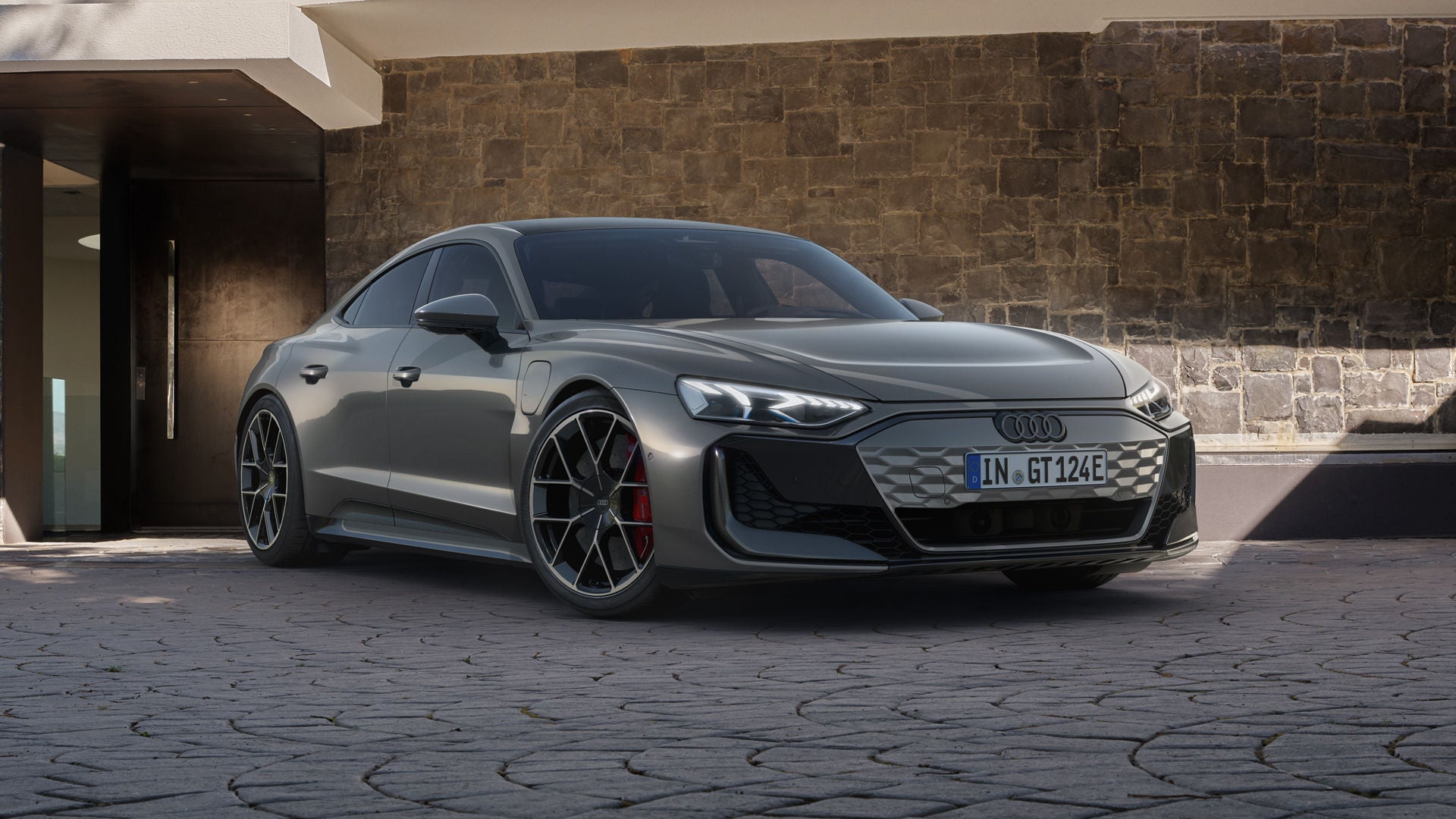 Der Audi RS e-tron GT schräg von vorn auf einem gepflasterten Parkplatz, im Hintergrund ein modernes Wohngebäude.