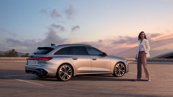 Junge Frau in Business-Look neben ihrem Audi A5 Avant auf einem Parkdeck bei tief stehender Sonne.