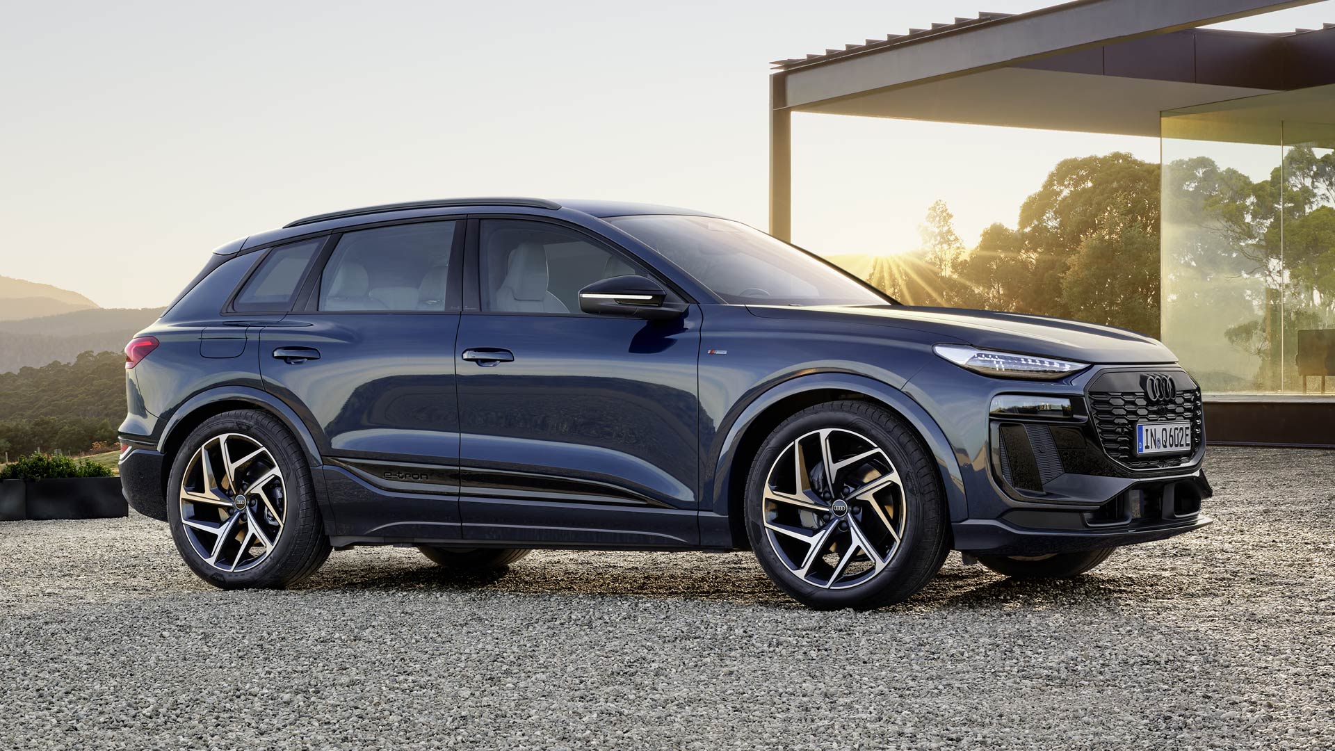 Q6 suv e-tron står på sidan på grusområdet framför ett modernt hus, upplyst av den nedgående solen.