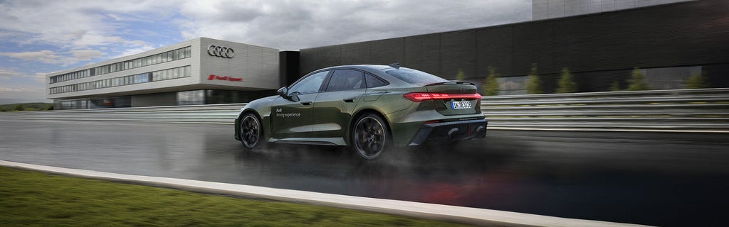 Grüne Audi RS 5 Limousine fährt auf nasser Strecke vor Audi-Gebäude, Rücklichter leuchten.