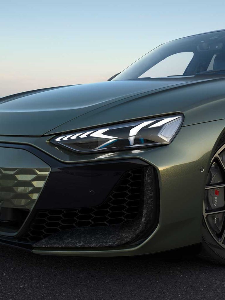 Nahaufnahme des Frontscheinwerfers und Kühlergrills eines grünen Audi RS e-tron GT performance mit Stadtblick im Hintergrund.