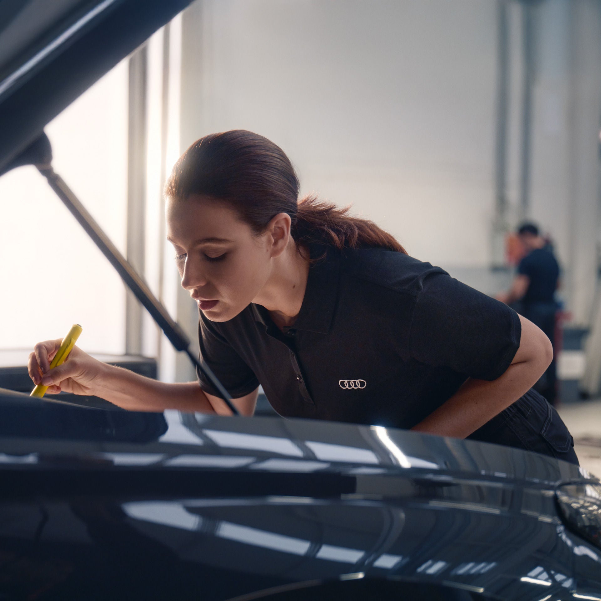 Eine Servicemitarbeiterin in schwarzem Audi-Poloshirt beugt sich über den geöffneten Motorraum eines Fahrzeugs und prüft mit einem gelben Messstab den Ölstand. Im Hintergrund ist eine Werkstattumgebung mit einer weiteren Person unscharf erkennbar.