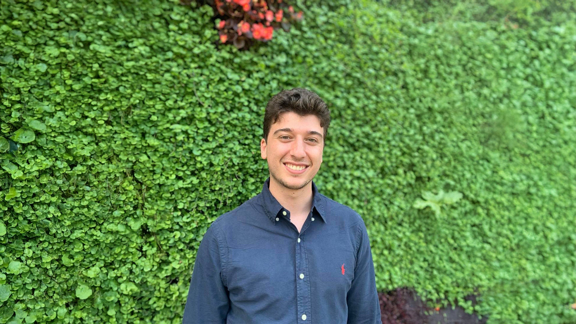 Junger Mann im dunkelblauen Hemd, lächelnd, vor grüner Pflanzenwand mit roten Blüten.