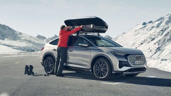 Person in roter Jacke verstaut Skier in Dachbox eines silbernen Audi vor verschneiten Bergen.