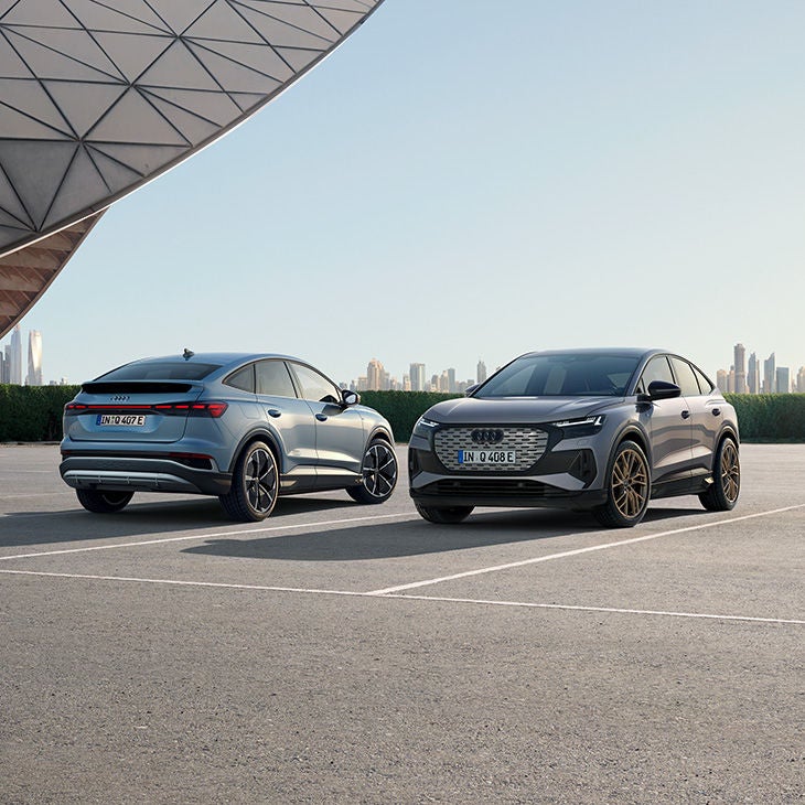Zwei Audi Q4 e-tron Modelle parken auf einem offenen Platz vor moderner Architektur und Skyline im Hintergrund.