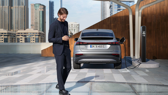 En mann holder en smarttelefon i hånden, en Audi lader ved en ladestasjon i bakgrunnen.