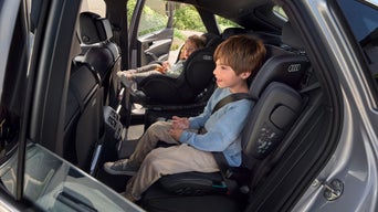 Zwei Kinder sitzen in Audi-Kindersitzen auf dem Rücksitz, eines lacht, das andere blickt ruhig nach vorn.