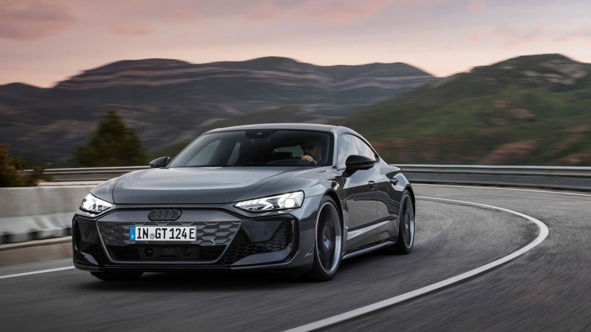 Grauer Audi e-tron GT fährt sportlich durch eine Kurve in bergiger Landschaft bei Sonnenuntergang.