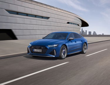 A blue sedan driving on a road with modern architecture and city skyline in the background.