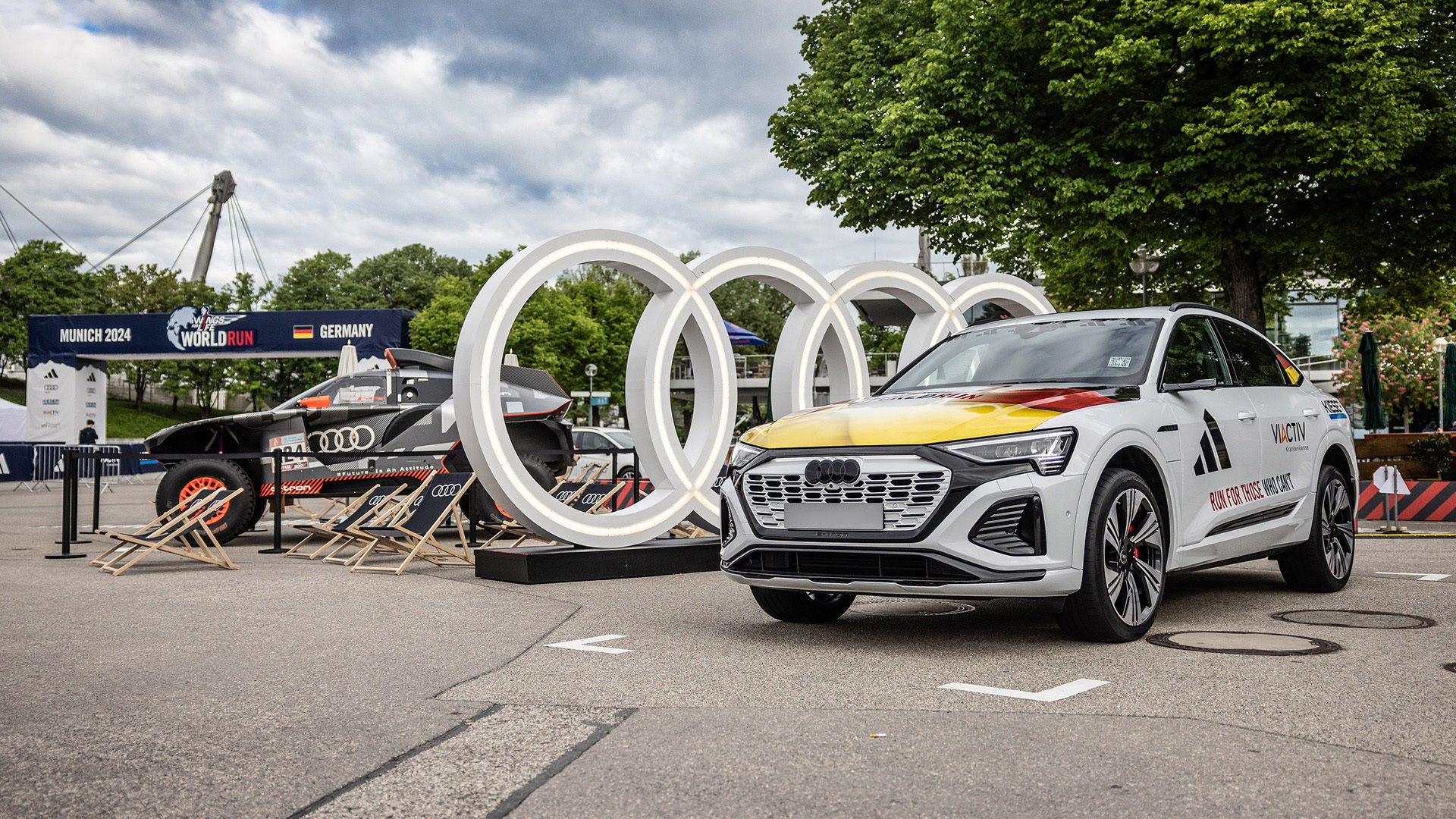 Weißer Audi Q8 e-tron 55 quattro mit Deutschlanddesign vor Audi Ringen; links ein ehemaliges Rallye-Dakar-Rennfahrzeug.