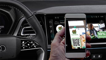 Smartphone mit QR-Code wird vor einem Infotainmentdisplay mit Werbeanzeige im Fahrzeug gehalten.