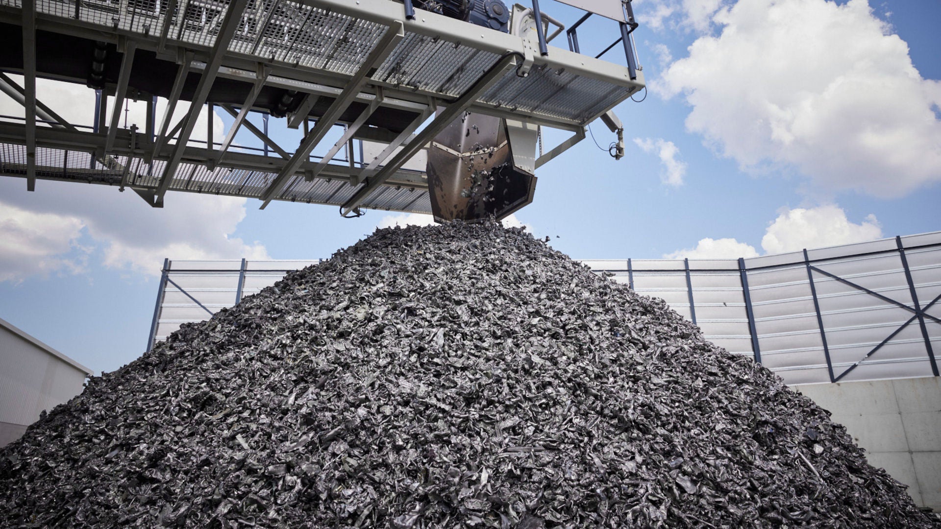 An excavator arm drops recycled materials onto a large pile of rubble at an outdoor facility beneath a blue sky. The parts are taken from end-of-life vehicles and reused for the recovery concept.