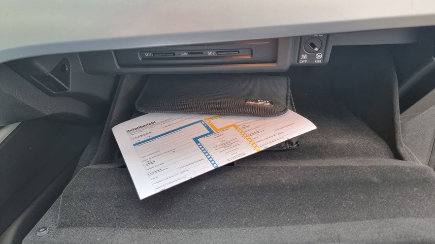Unfallbericht und schwarze Audi-Mappe liegen im geöffneten Handschuhfach eines Fahrzeugs.