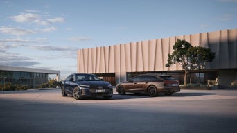Deux Audi A6, une berline bleue et une Avant brune, stationnées devant un bâtiment moderne aux lignes architecturales épurées, dans un environnement urbain contemporain.