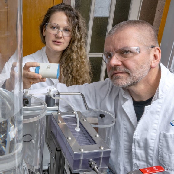 Eine Mann und eine Frau in Laborkitteln und Schutzbrillen betrachten in einem Labor einen Zylinder mit Flüssigkeit.