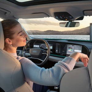 The interior of the Q6 SUV e-tron, viewed from the rear seat. A young woman is sitting in the driver's seat, leaning back with her eyes closed. Through the windshield, you can see the ocean and mountains.