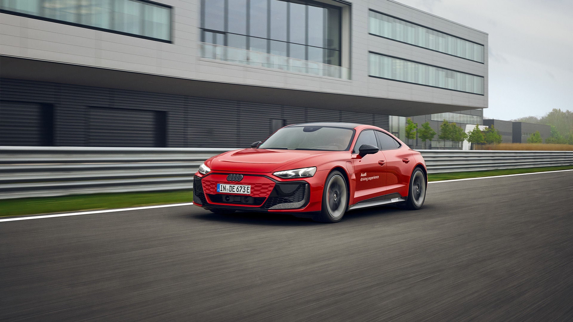 A red Audi e-tron GT drives along a race track in front of a modern building with a glass façade.