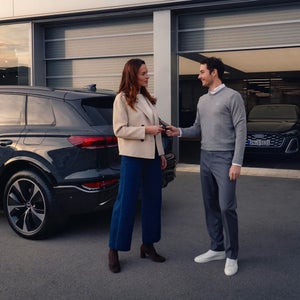 A woman in a cream blazer hands a car key to a man in a grey sweater outside a modern vehicle, with another car visible in the background.