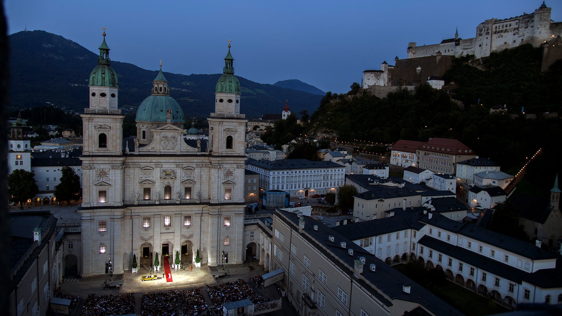 Salzburger Dom und Festung bei Dämmerung mit beleuchteter Bühne und Publikum auf dem Domplatz.