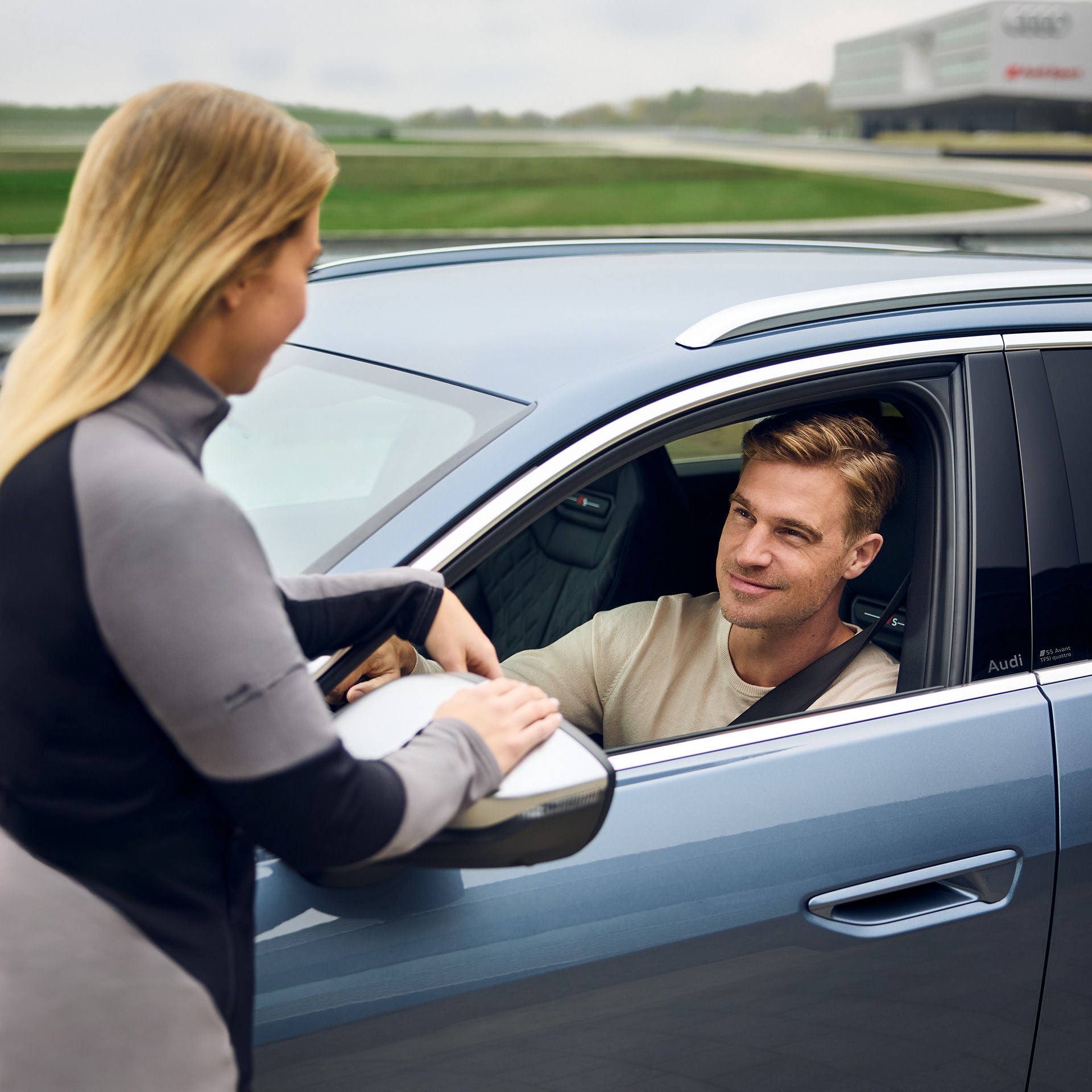 Mann im Audi unterhält sich mit einer blonden Frau außerhalb des Fahrzeugs auf einer Rennstrecke.