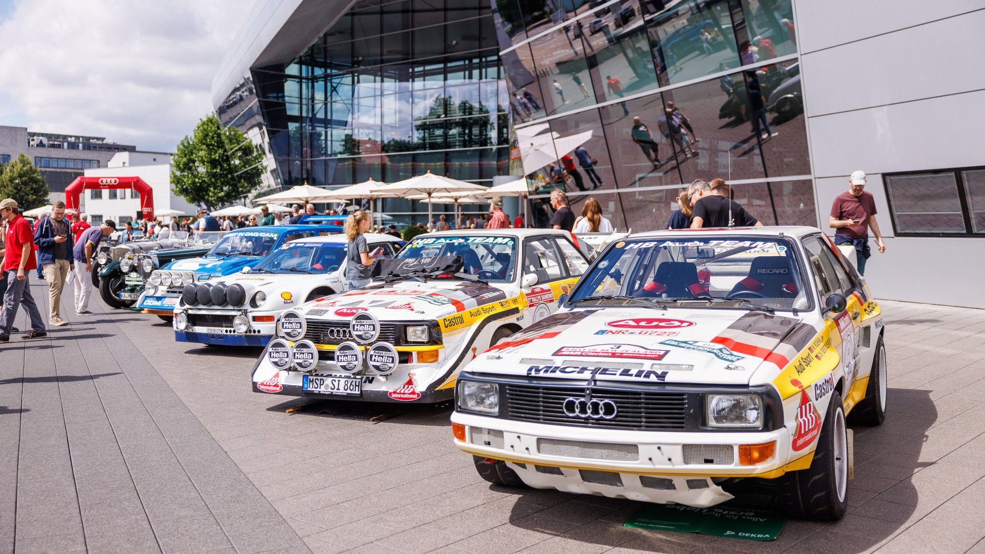 Mehrere historische Audi-Rallyefahrzeuge in Weiß-Gelb mit Sponsorenlogos vor dem Audi Forum Neckarsulm.