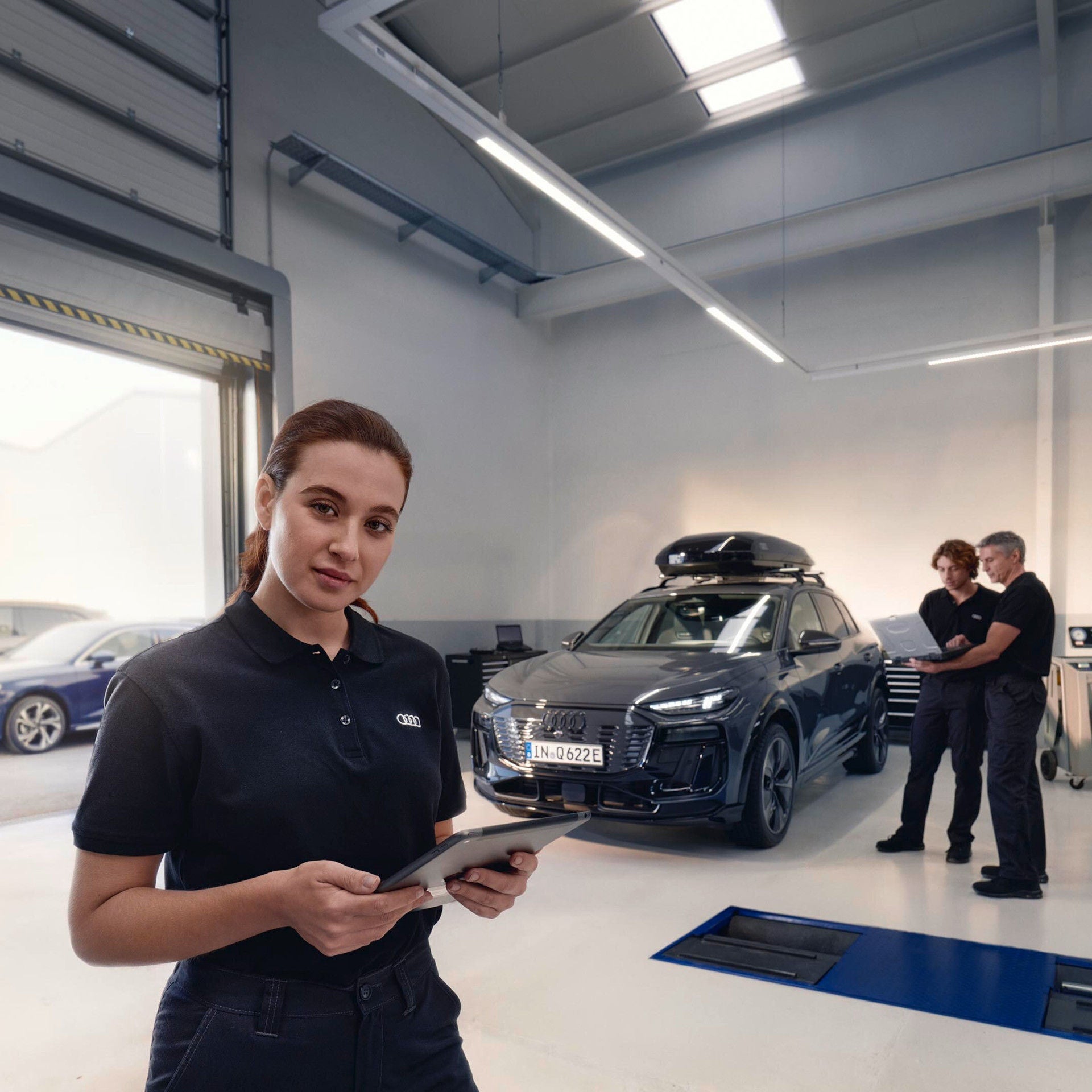 Mitarbeiterin hält ein Tablet, während zwei Kollegen neben einem Auto arbeiten.