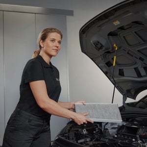 A technician in a black polo shirt holds an air filter above a car engine, ready for maintenance in a well-lit garage.