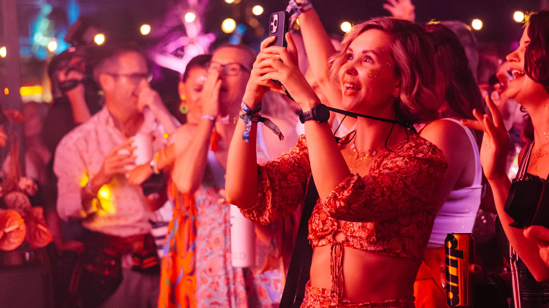 A woman in a floral outfit films the stage on her phone, smiling, surrounded by a dancing crowd lit in warm red and pink tones. A woman in a floral outfit films the stage on her phone, smiling, surrounded by a dancing crowd lit in warm red and pink tones.