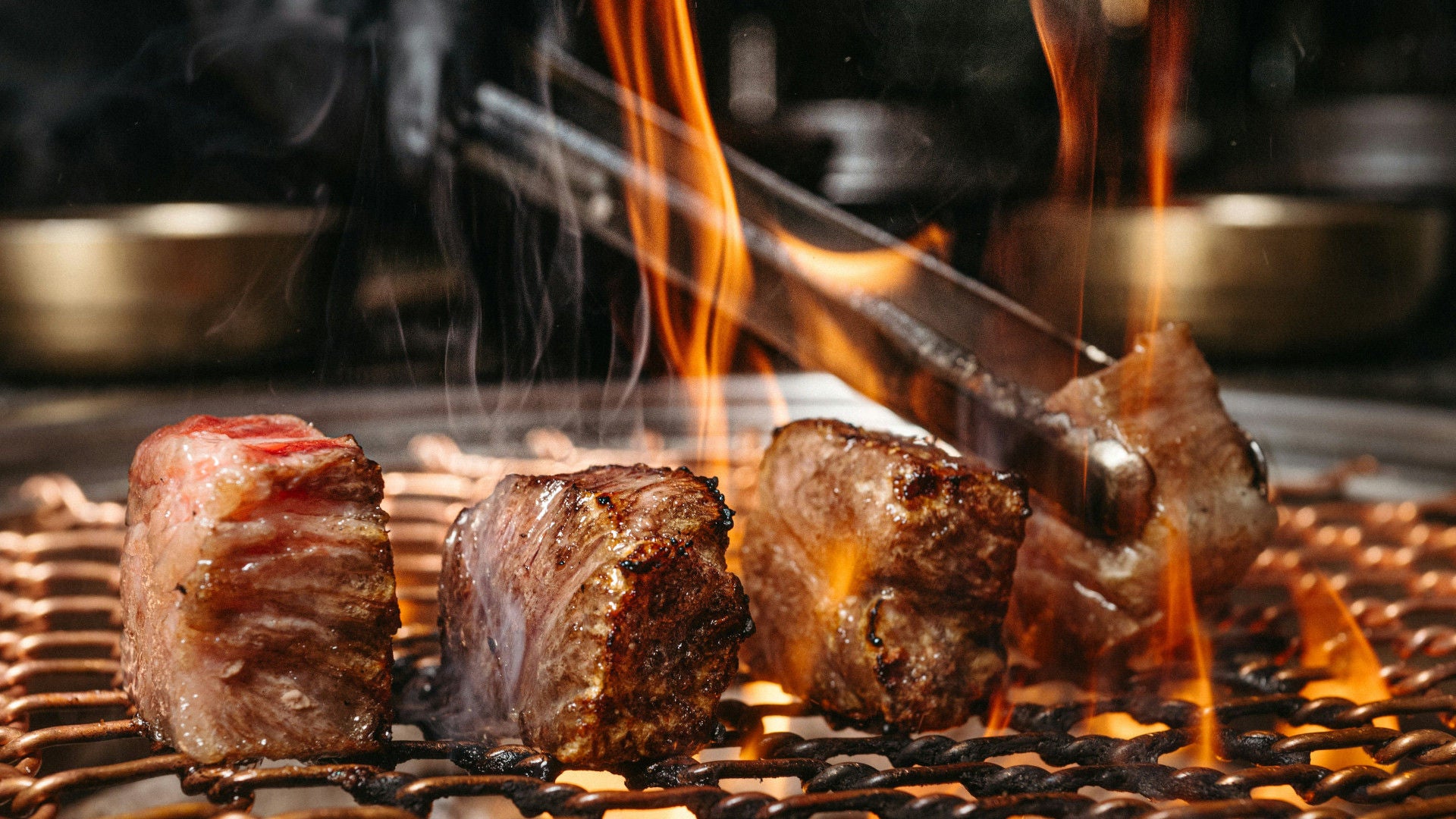 Drei Stücke Fleisch braten auf einem Grillrost; Flammen und Rauch steigen auf, eine Grillzange hält ein Stück.