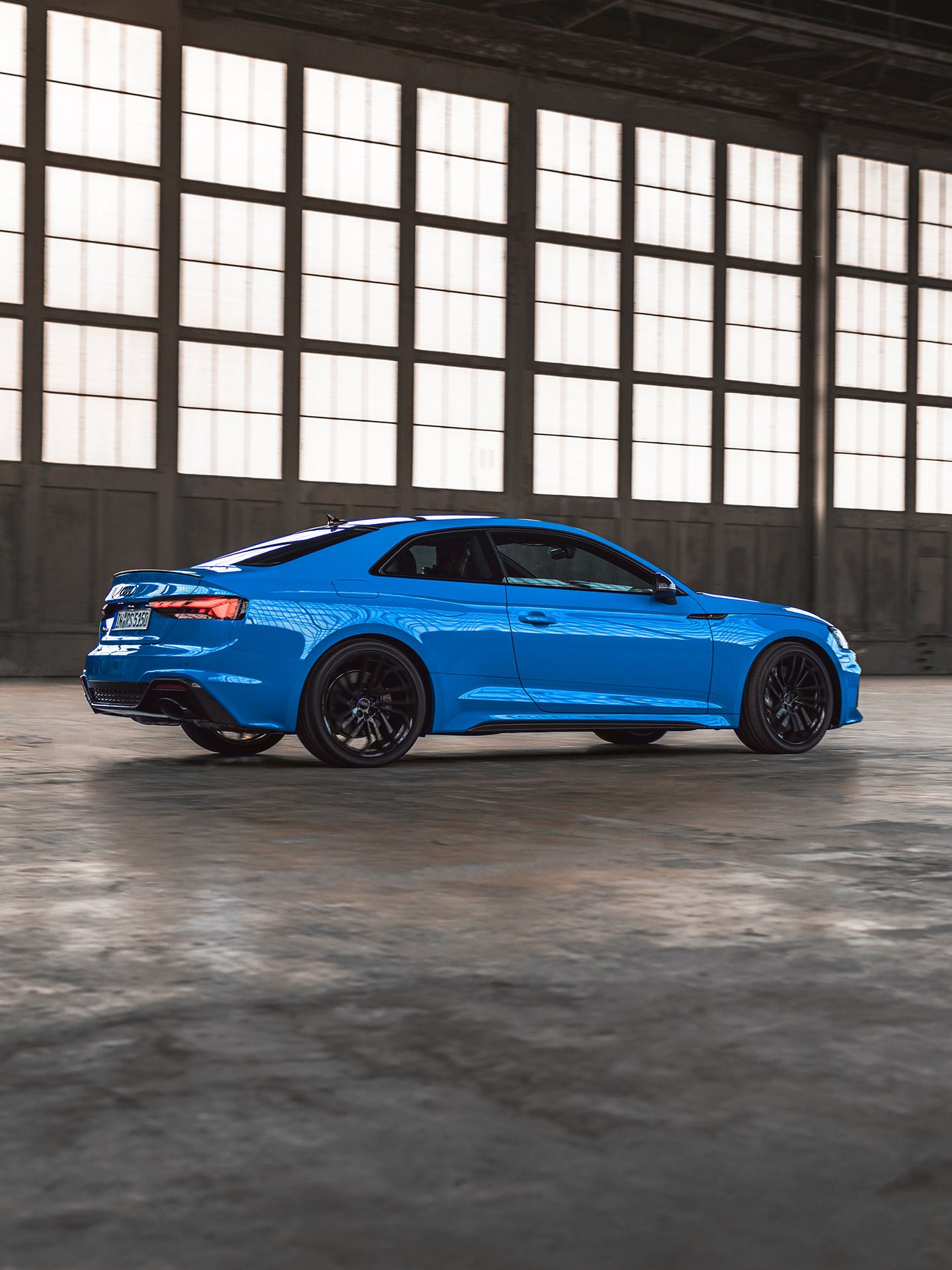 Blauer Audi RS 5 fährt durch eine helle Industriehalle mit großen Fensterfronten, Seitenansicht des sportlichen Coupés.