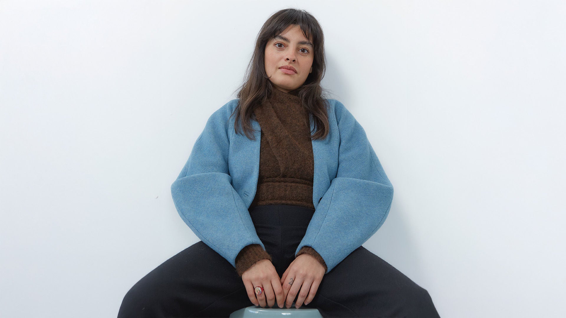 Frau mit mittellangen dunklen Haaren im braunen Pullover und blauer Jacke, vor weißem Hintergrund sitzend.